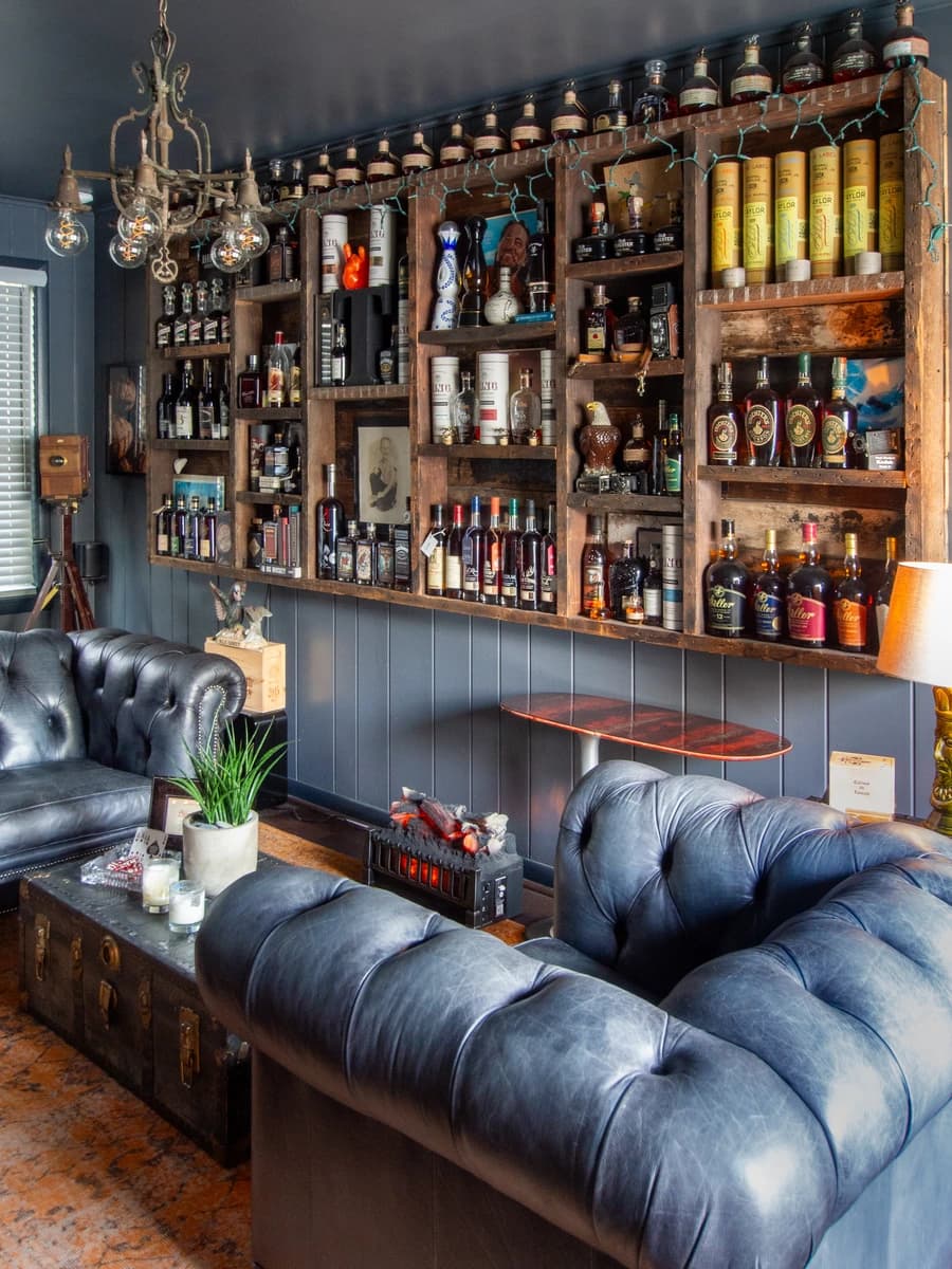 Large leather sofa against wall, rustic shelves filled with bottles, wooden bench at bottom.