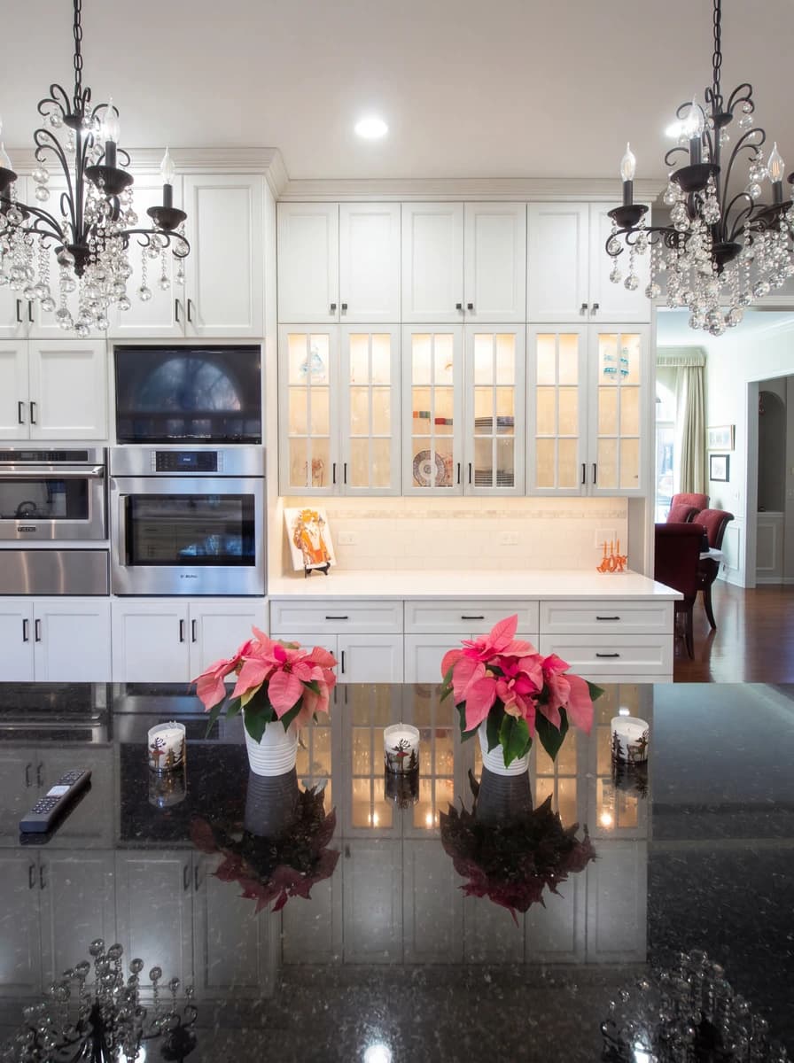Elegant kitchen with white cabinetry, marble countertops, chandeliers, and potted plants.