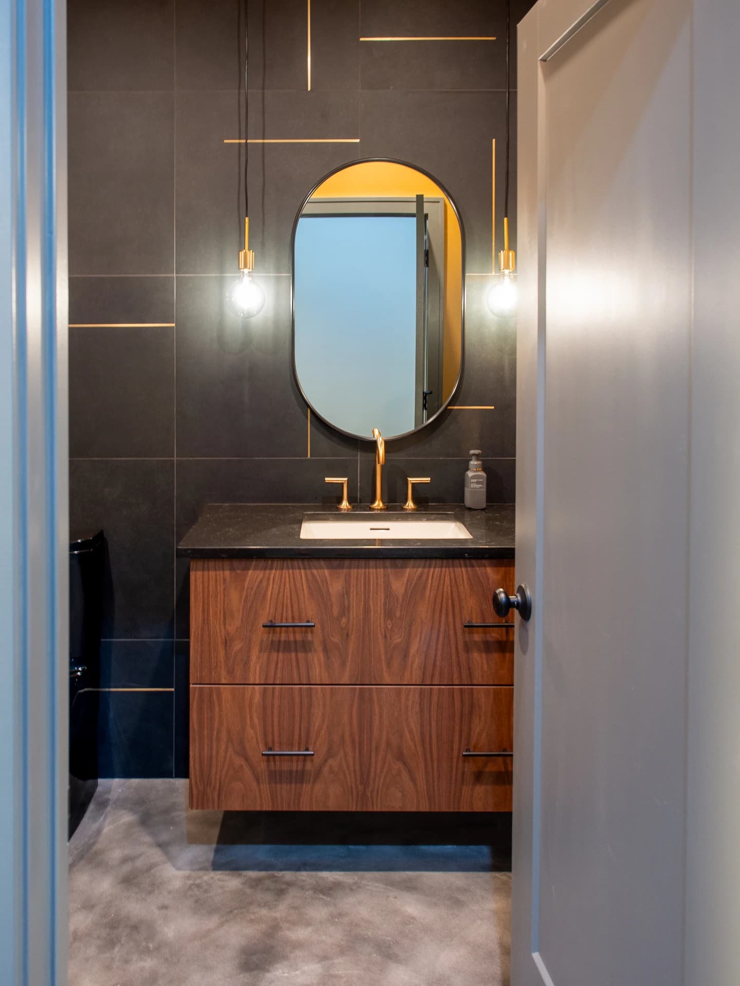 Modern bathroom with black and grey walls, brown vanity, and gold accents.