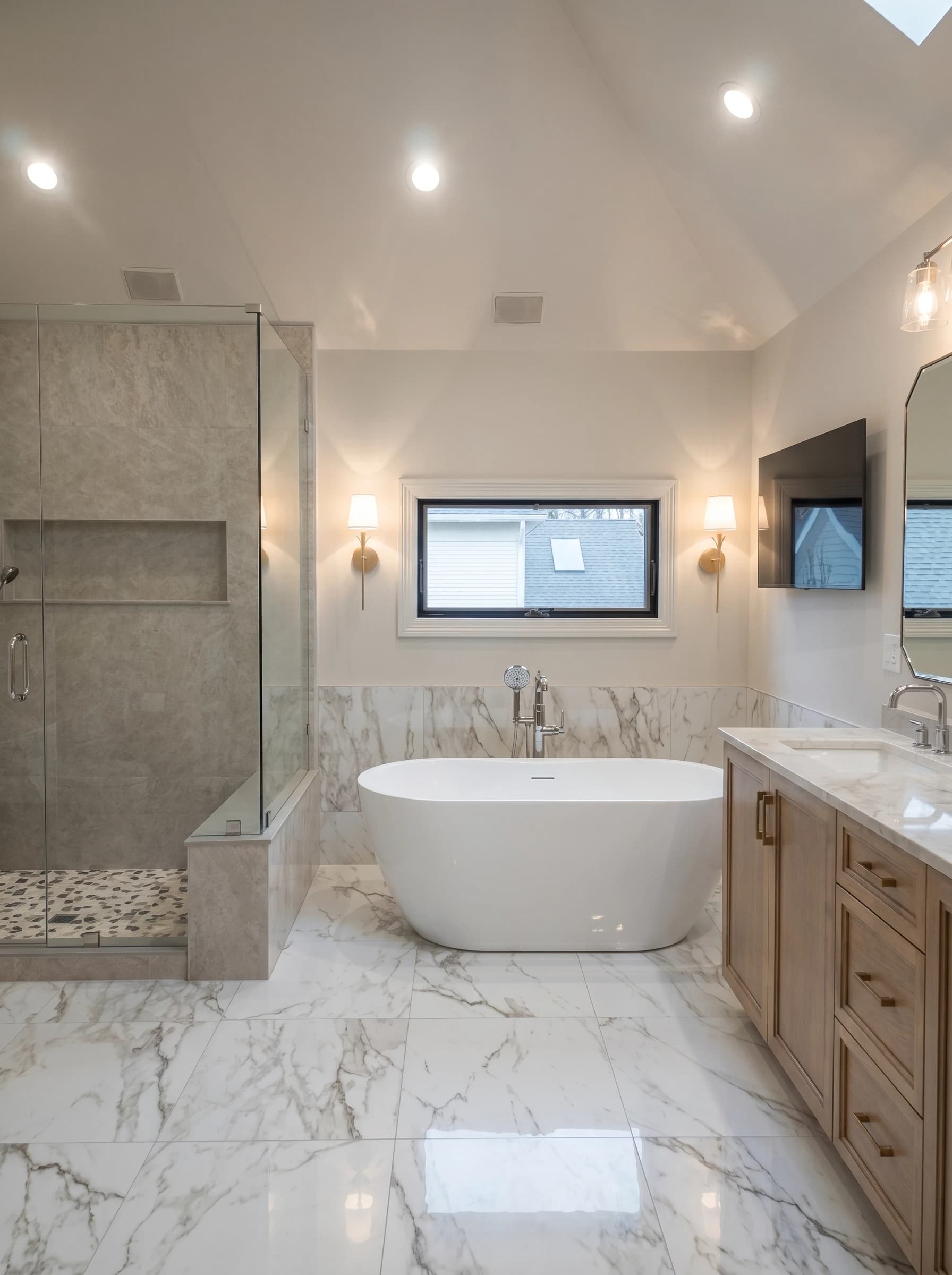 Modern bathroom with white marble floors and walls, wooden cabinetry, white sink, and large bathtub.