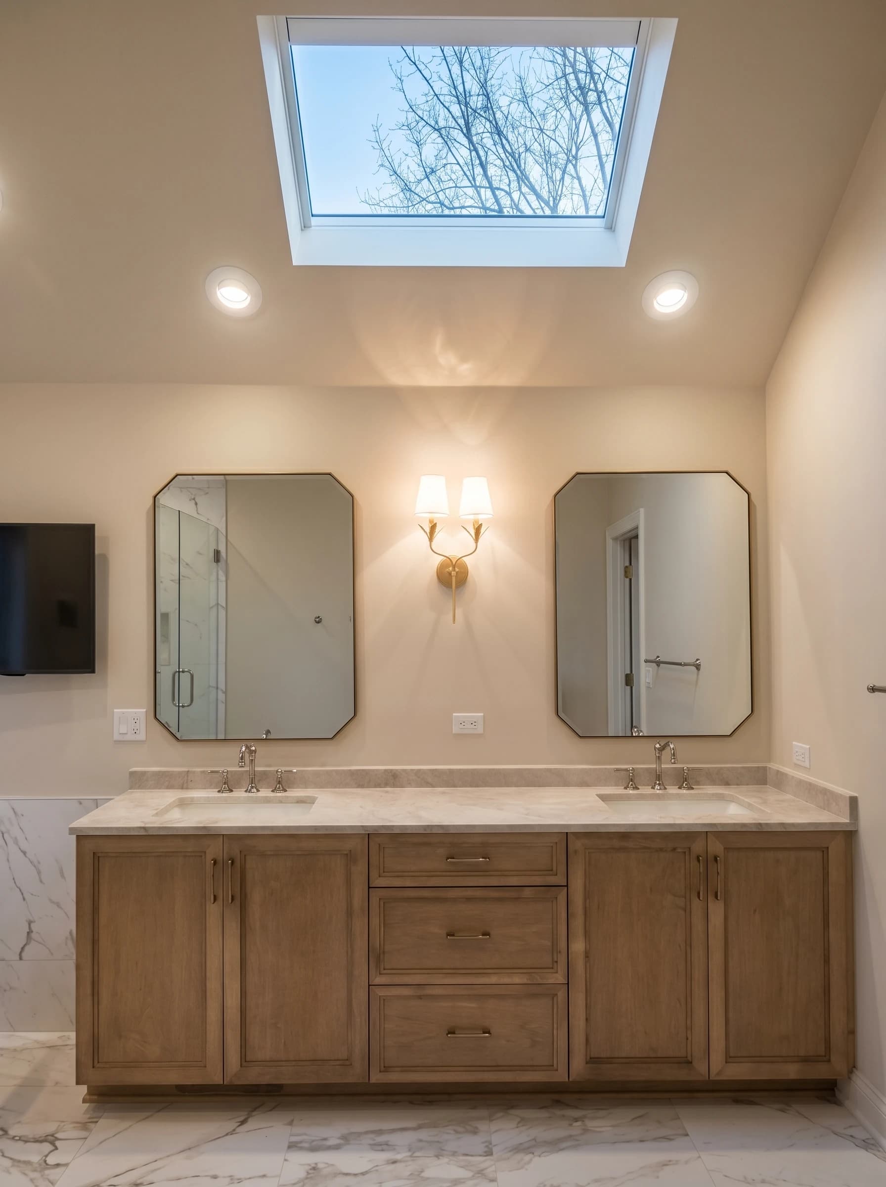 Clean, modern bathroom with sleek wood cabinets, white marble counters, large mirror, and skylight.