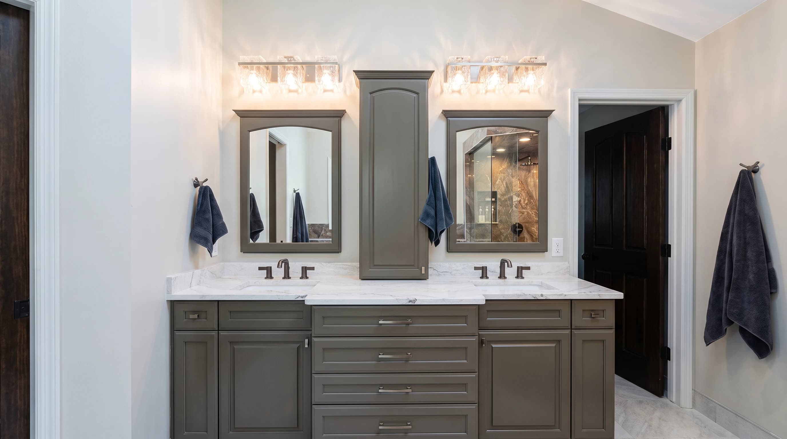 Symmetrical olive green double vanity with white marble tops, bronze fixtures, framed mirrors, and flanking wood doors.
