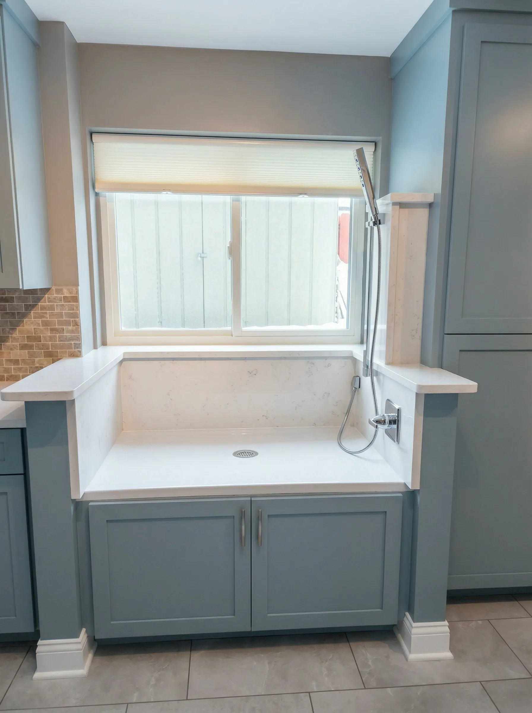 Blue custom dog washing station cabinets and sink, large window above white countertop, grey tile flooring.