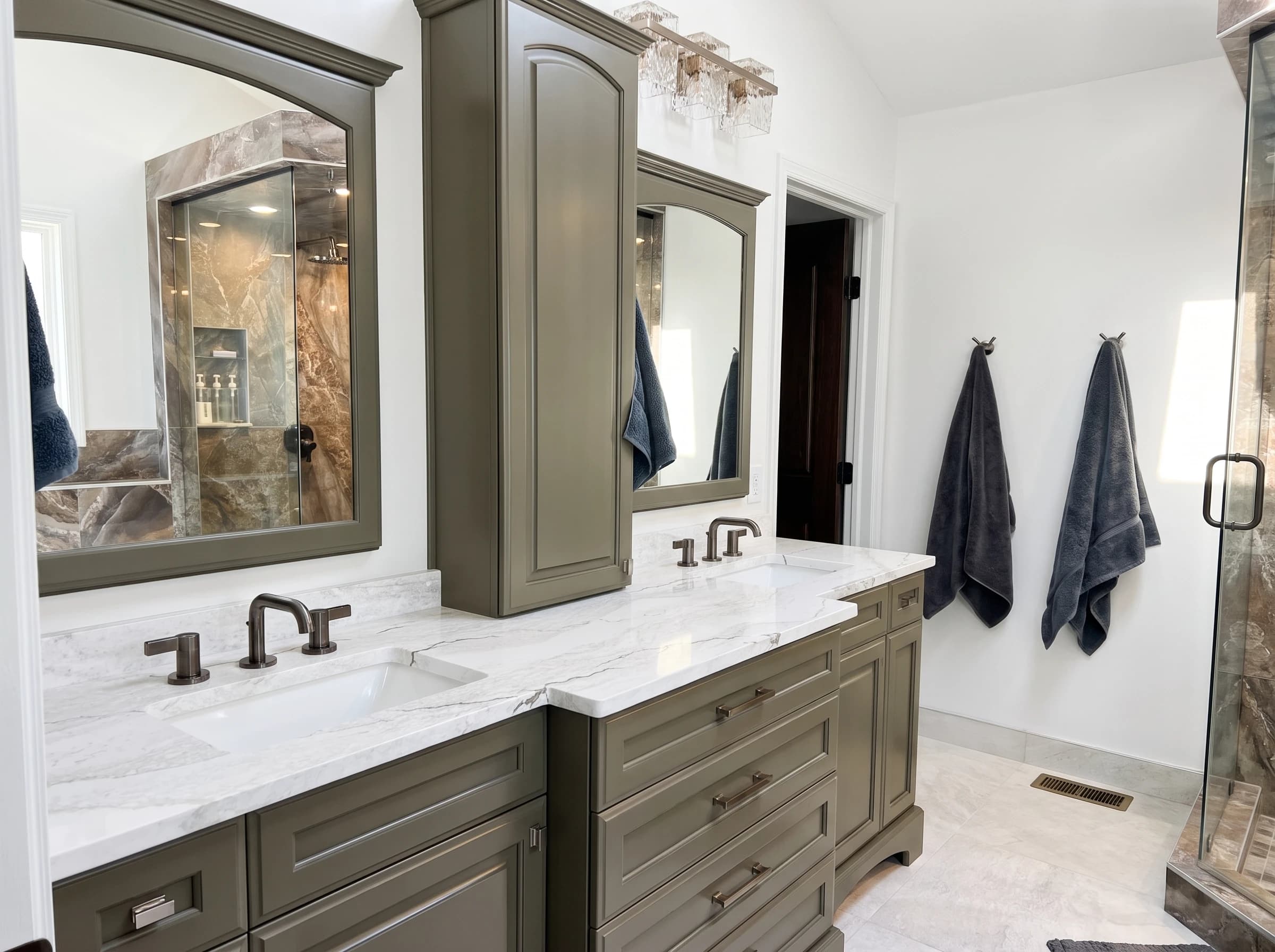 Primary bathroom with olive green double vanity, white marble countertops, bronze faucets, arched mirror, and tall linen cabinet.